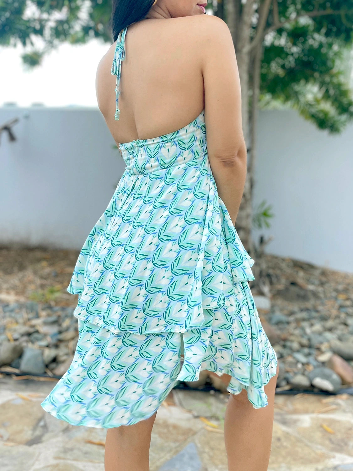 Back view of a woman modeling a backless summer dress showing the open back detail and the tie closure at the neck. The asymmetrical double-layer skirt is also visible.