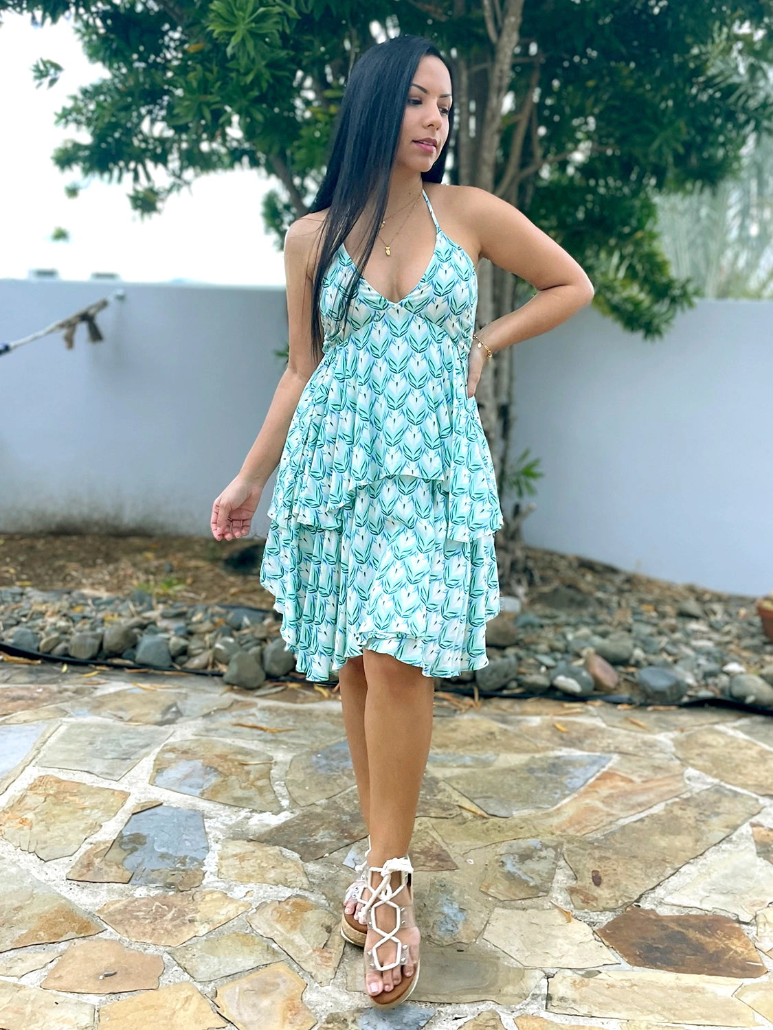 Woman in a short, flowy aqua and white tropical print halter neck dress with an open back. She is wearing white gladiator-style sandals and posing outdoors on a stone walkway.