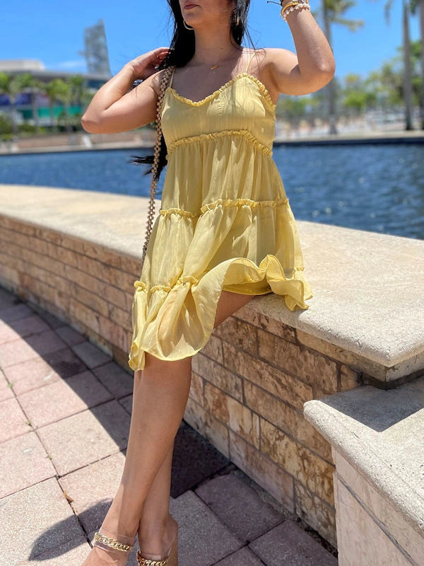 Woman sitting on a ledge by the water, wearing a flowy yellow sundress with tiered ruffles and delicate straps.