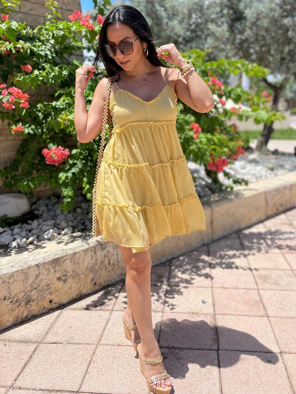 Woman wearing a short yellow summer dress with tiered layers and thin straps, accessorized with sunglasses and bracelets.